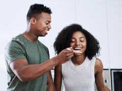 A young Black couple tastes what they're making in the kitchen with a wooden spoon.
