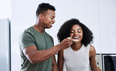 A young Black couple tastes what they're making in the kitchen with a wooden spoon.