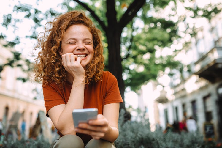 A young woman laughs while sitting outside in the summer and socializing through a video chat.