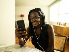 A young woman sits at her kitchen table and video chats on her phone while wearing headphones.