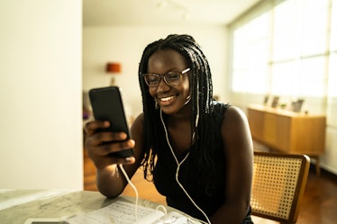 A young woman sits at her kitchen table and video chats on her phone while wearing headphones.