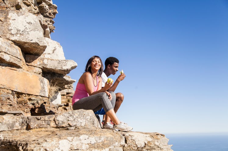 A young couple sits at the top of a mountain after a summer hike and eats a vegan snack.