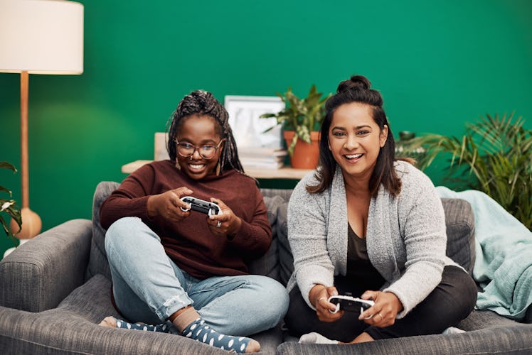 Two young women sit on a couch in front of a bright green wall and play video games.