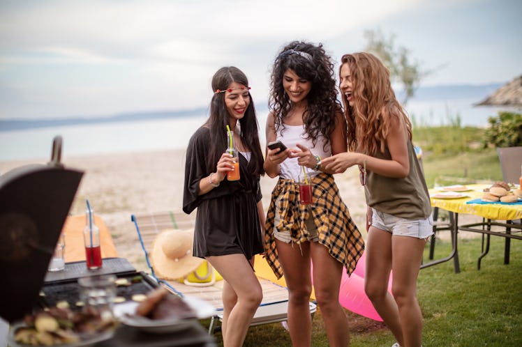 Three friends hang out at a barbecue outside, while laughing at something on their phones. 