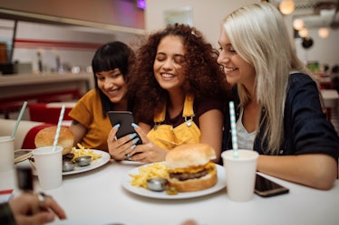 Three happy friends look at their phone, while sitting at a diner with burgers in front of them. 