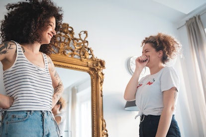 Two young women laugh while trying on summer outfits and posing in a vintage mirror.
