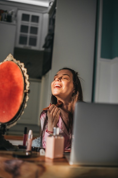 A young woman smiles while applying makeup and looking at herself in a small, vintage mirror.