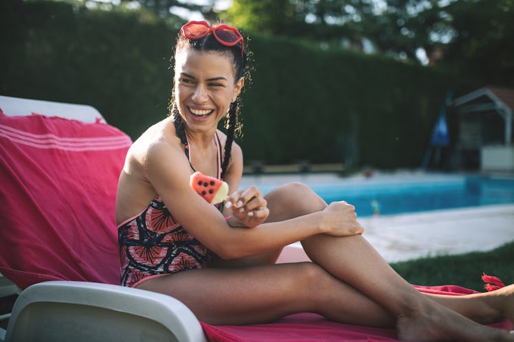 A happy woman sits on a lounge chair with a watermelon popsicle in her backyard during the summer. 