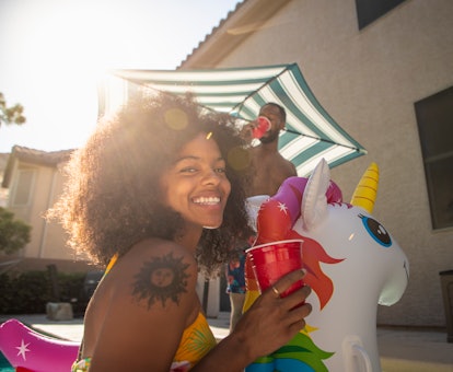 A happy woman sits on a unicorn pool float, while hanging in her backyard during the summer. 