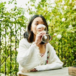 A woman sitting outside and drinking wine at a table surrounded by greenery