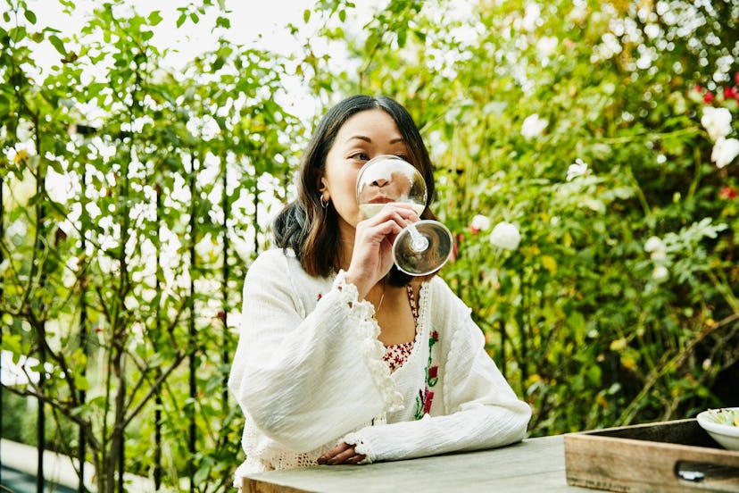 A woman sitting outside and drinking wine at a table surrounded by greenery