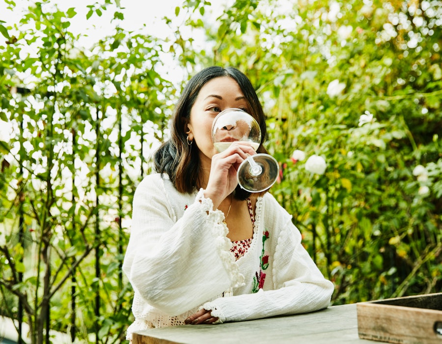 A woman sitting outside and drinking wine at a table surrounded by greenery