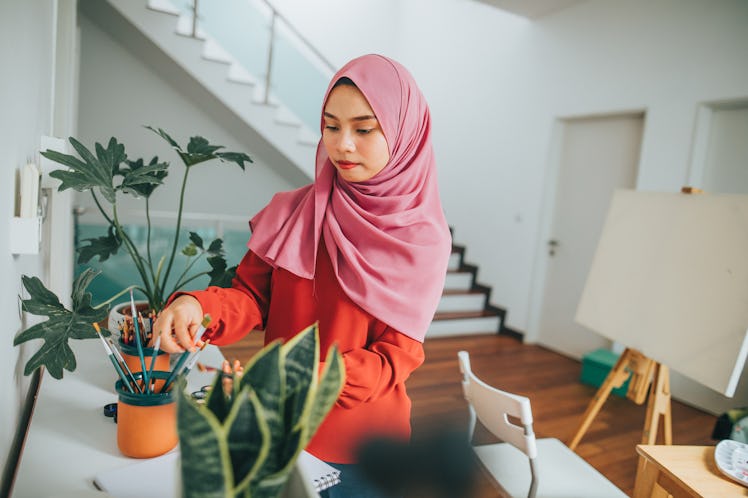 A young woman reaches for paintbrushes before working on a canvas that's set up in her living room.