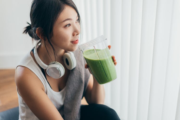 A young Asian woman sips on a green smoothie after a workout, while sitting on a yoga mat.