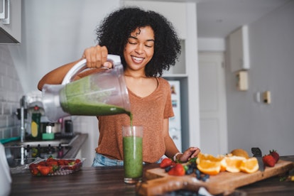 A young Black woman pours a green smoothie into a glass after cutting up fresh fruit in her kitchen.