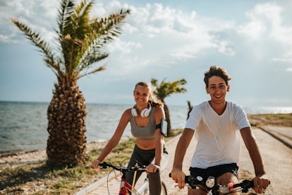 A young couple goes biking next to the ocean at sunset, and smiles for the camera.