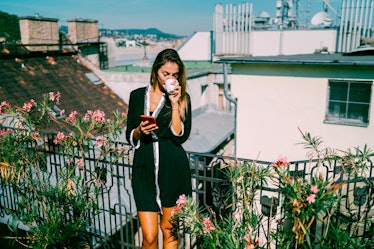 A young woman sips on coffee while standing on her balcony that's covered with plants.