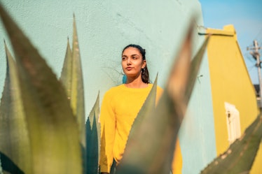 A young woman poses against a teal wall, while also standing behind a large succulent.
