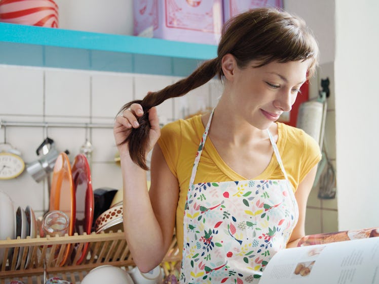 A woman twirls her hair, while looking through a cookbook in her kitchen. 