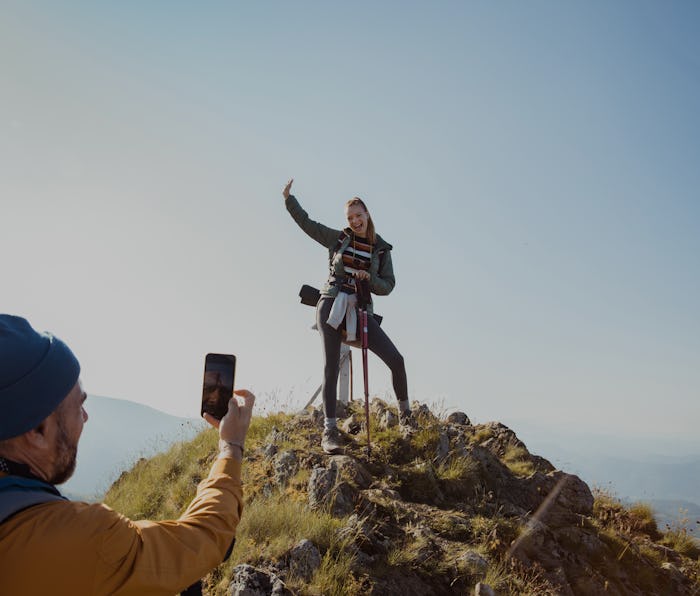 A woman is smiling and standing atop a hill while posing for a photo a man is taking of her from afa...