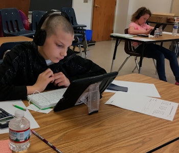 A young student can be seen looking at their tablet in a classroom. They have headphones on.