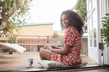 A happy woman sits on a pillow with a mug next to her, while outside on her patio. 