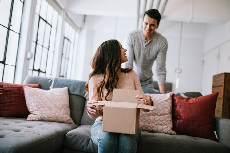 A young couple smiles while opening up a gift box on their couch.