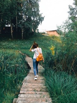 A young woman walks down a boardwalk in a lush field with a backpack on.