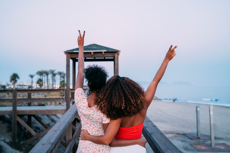 Two Black girls pose on a boardwalk near the beach, and throw peace signs up in the air.