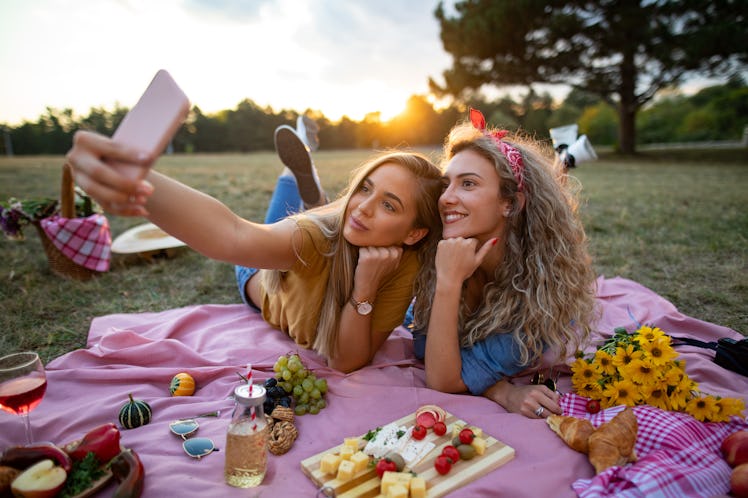 Two friends take a selfie during their wine and cheese board picnic outside.
