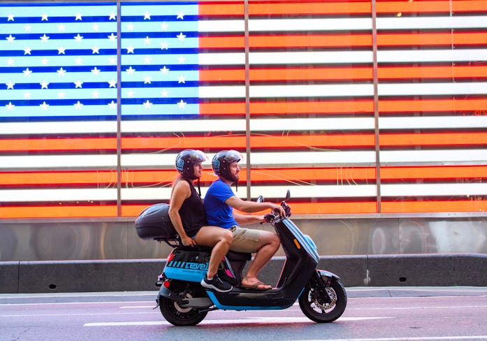 A man and woman can be seen on a Revel moped in front of the United States flag.