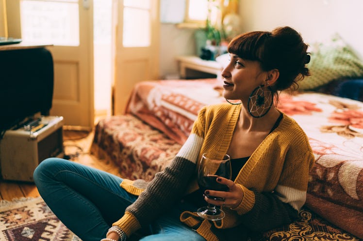 A woman wearing a yellow cardigan, sits on her bedroom floor, holding a glass of wine.