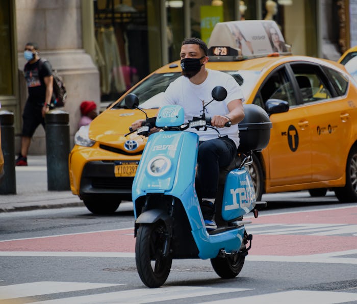 A man wearing a white shirt can be seen riding a Revel moped. Behind him there is a New York taxi. 