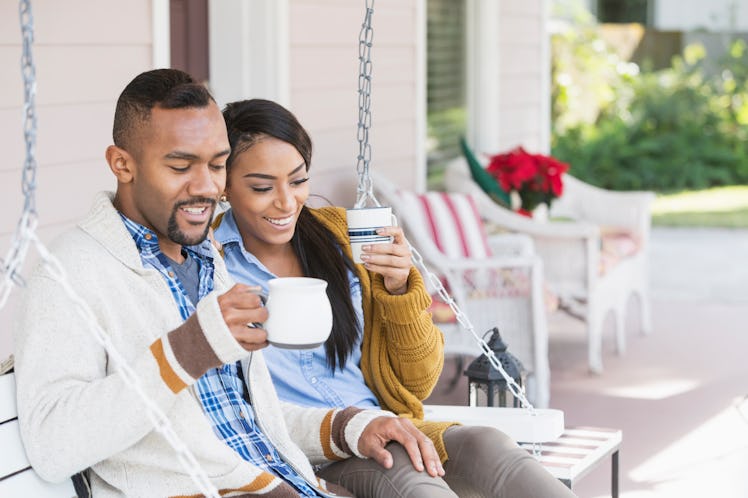 A couple wearing cardigans, sit on their front porch, holding mugs. 