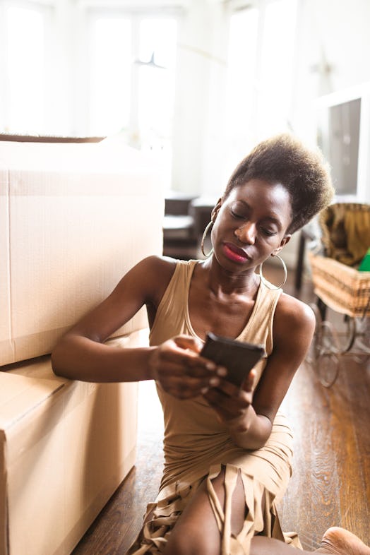 A young Black woman sits next to cardboard boxes on her apartment floor and texts on her phone.
