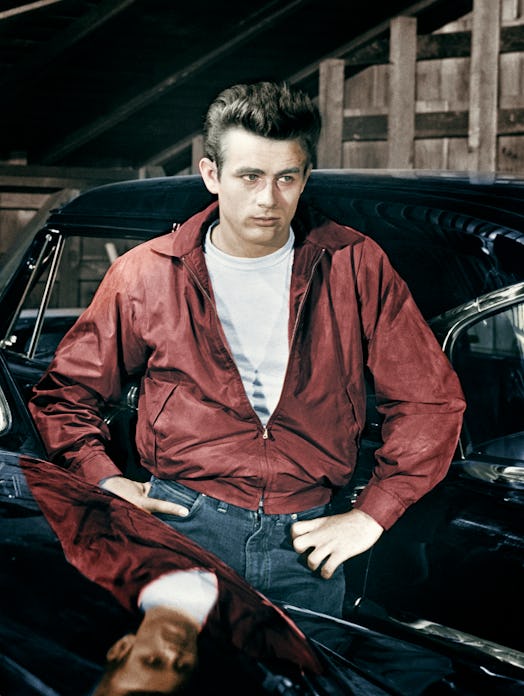James Dean posing between two cars in a red jacket, white shirt and blue denim jeans