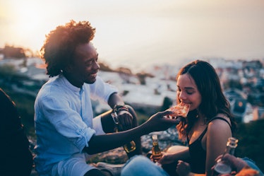 A young couple drinks beer and eats pizza while sitting outside on a blanket at sunset.