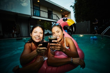 A young lesbian couple hangs out in a pool at night with glasses wine and a flamingo pool float.