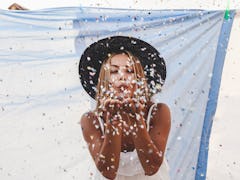 A young woman stands in front of a blue sheet on the beach and blows confetti at a camera.