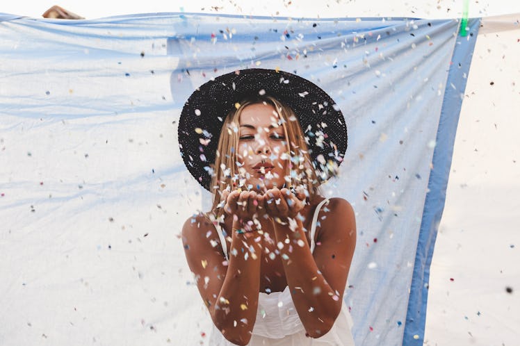 A young woman stands in front of a blue sheet on the beach and blows confetti at a camera.