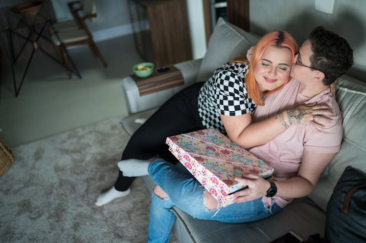 A couple sitting on a couch, embracing while holding a gift wrapped in colorful paper, in a cozy liv...