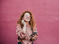 A young woman with red hair stands in front of a hot pink wall with her phone and laughs.