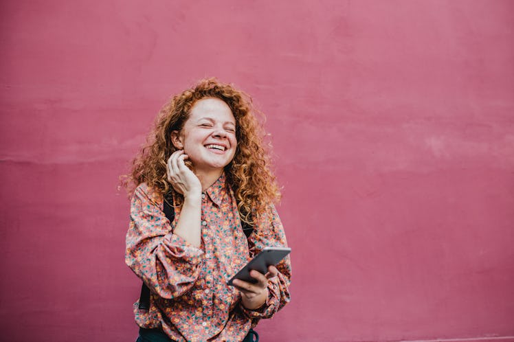 A young woman with red hair stands in front of a hot pink wall with her phone and laughs.