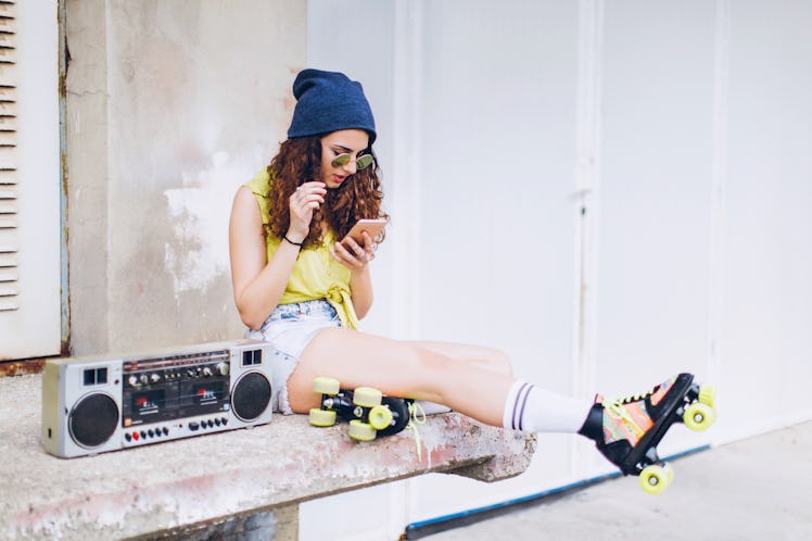 A girl with roller skates on, sitting next to a boombox, is looking at her phone. 