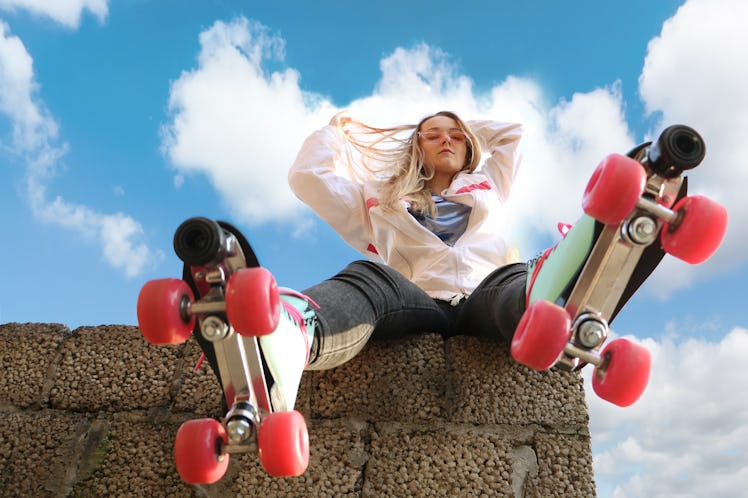 A young woman sits on a concrete ledge and poses with her pink, blue, and white rollerblades.