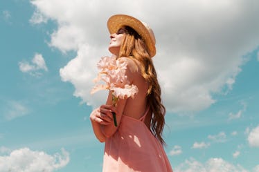 A young woman with long, red hair holds pink flowers and looks up at the sky on a sunny day.