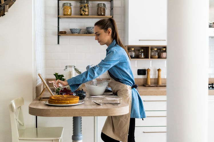 A young woman bakes a fruity cake in her kitchen while wearing an apron and scrolling on her tablet.