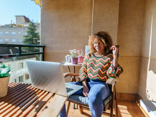 A young Black woman sits at a table on her patio on a sunny day and works remotely.