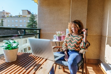 A young Black woman sits at a table on her patio on a sunny day and works remotely.