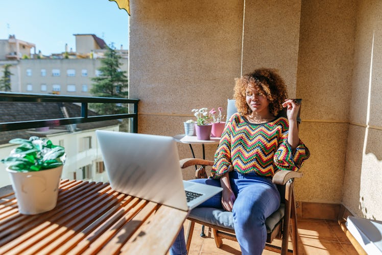 A young Black woman sits at a table on her patio on a sunny day and works remotely.